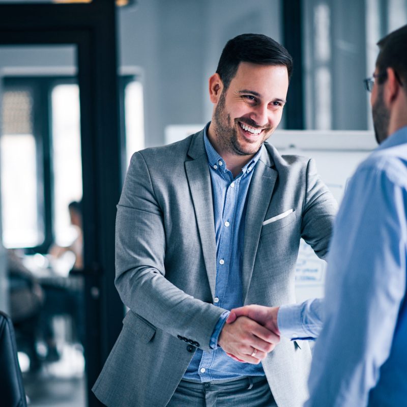 Two smiling businessmen shaking hands while standing in an office.